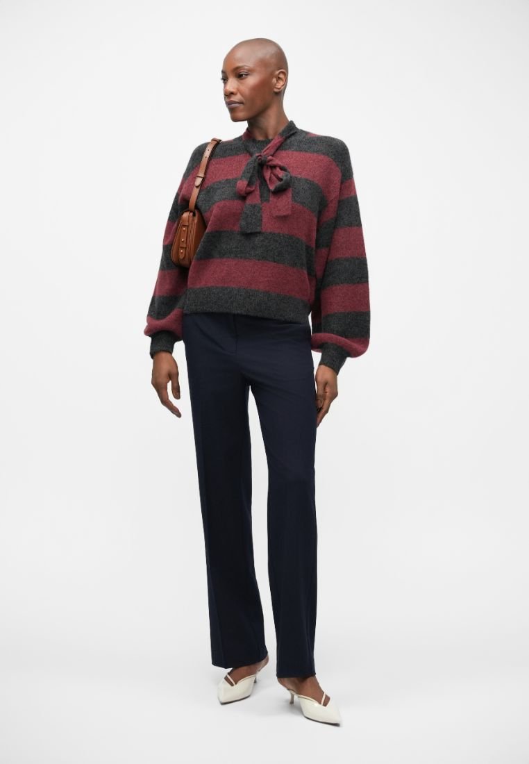 Red and gray striped, cropped sweater with bow detail at the neckline. Paired with navy trousers and white pointed-toe shoes. Small brown handbag.