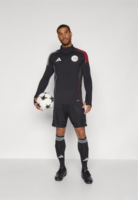Black athletic track jacket with red and white stripes, logo on chest, paired with black shorts, socks with white stripes, and black cleats. Holding soccer ball.