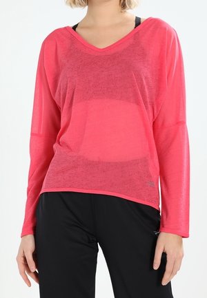 Vrouw draagt een doorschijnend roze top met lange mouwen en V-hals over een zwarte sportbeha en zwarte broek.