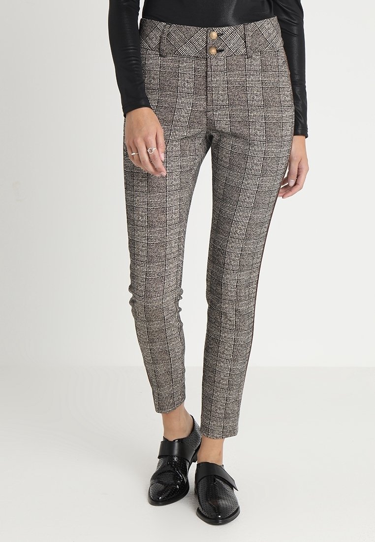 Femme portant un pantalon slim à carreaux taille haute avec double boutons devant, chaussures noires et un haut noir à manches longues, debout sur un fond clair.
