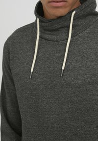 Mørkegrå sweatshirt med høj hals, der har lysebeige snore og et blødt, tekstureret stof. Nærbillede af halsåbningen.