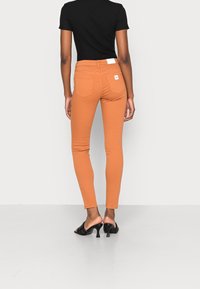 Jeans ajustados en un color óxido, hechos de denim con una textura suave. Presentan dos bolsillos traseros y una etiqueta en la cinturilla. Combinados con mules negras de tacón.