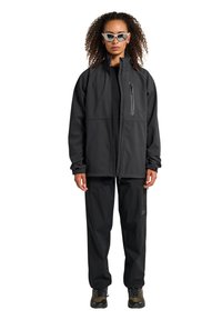 Schwarze isolierte Jacke mit hohem Kragen, Reißverschlusstasche, kombiniert mit schwarzen Hosen mit dezentem Muster und grünen Wanderschuhen. Elegante Passform.