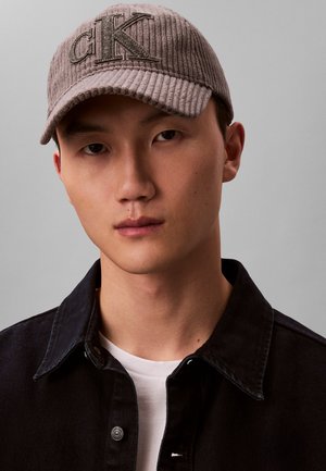 Giovane uomo con un cappellino grigio a coste CK, giacca di denim nera sopra una maglietta bianca, su sfondo uniforme grigio chiaro.