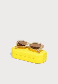 Óculos de sol com armação transparente e lentes castanhas, apresentando um detalhe em rosa. Colocados sobre uma caixa amarela texturizada de forma retangular.