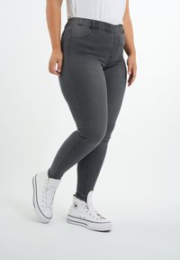 Jean skinny gris avec une taille élastique, présentant une texture lisse et des coutures contrastées. Associé à des baskets blanches montantes.