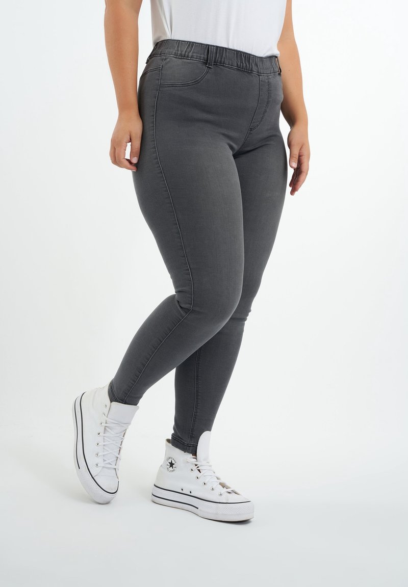 Jean skinny gris avec une taille élastique, présentant une texture lisse et des coutures contrastées. Associé à des baskets blanches montantes.