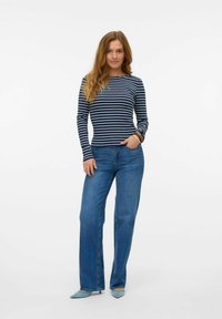 Marinblå och vitrandig långärmad topp i kombination med blå vida jeans. Modellen står med en hand i fickan, mot en ljus bakgrund.