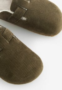Sabots en velours côtelé marron avec un intérieur doux, présentant une lanière sur le dessus et un bout arrondi. Surface texturée avec des coutures visibles.