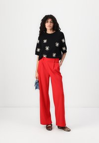 Pull noir avec broderie florale blanche, associé à un pantalon large rouge. Accessoirisé avec une pochette bleue et des chaussures à imprimé animal.