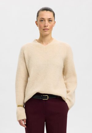 Frau mit zurückgebundenen Haaren, die einen beigen Strickpullover und dunkelweinrote Hose mit schwarzem Gürtel trägt, steht vor einem schlichten weißen Hintergrund.