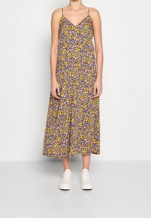 Mujer vistiendo un vestido floral sin mangas, largo hasta los tobillos, con flores amarillas y moradas, combinado con zapatillas blancas de cordones.