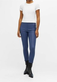 Haut blanc à manches courtes associé à un pantalon bleu ajusté. Le modèle porte des bottines noires. Tissu doux avec un léger éclat, design minimaliste.
