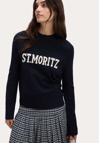 Granatowy sweter z białym napisem "ST. MORITZ". D�ługie rękawy, ściągacz na szyi i u dołu. W zestawie z czarno-białą spódnicą w kratę.