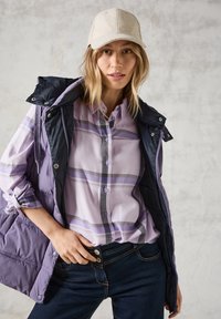 Chaqueta sin mangas acolchada en púrpura y azul marino sobre una camisa de cuadros lavanda con botones, combinada con jeans de color azul oscuro. Presenta una gorra de color claro.