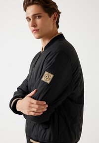 Jachetă bomber neagră cu textură matlasată, guler și manșete ribbate, având un patch cu logo bej pe mânecă.