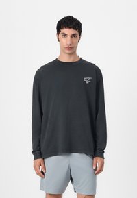 Black long-sleeve cotton t-shirt with a round neckline and logo detail on the chest. Paired with light gray shorts.