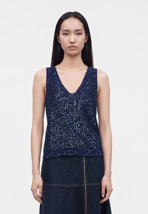Kobieta z długimi czarnymi włosami, ubrana w ciemnoniebieski top bez rękawów ozdobiony cekinami oraz ciemną spódnicę dżinsową z widocznymi szwami i zamkiem.