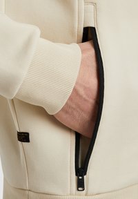 Sweat-shirt beige avec une poche zippée, des poignets côtelés et un tissu texturé. La poche possède une fermeture éclair noire contrastante.