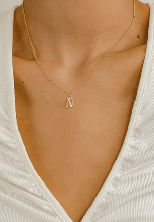 Collier doré présentant un pendentif rectangulaire avec la lettre "N" ornée de subtils strass, reposant sur un haut blanc texturé.
