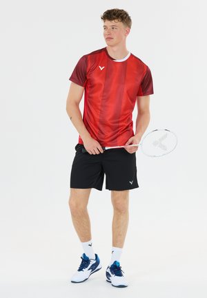 Ung mand iført rød sportstrøje og sorte shorts, der holder en badmintonketcher, stående på en hvid baggrund.