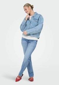 Mlada žena u svijetloplavoj traper jakni i trapericama, bijeloj majici, crvenim balerinkama s kopčama, stoji s prekriženim nogama i smiješi se.