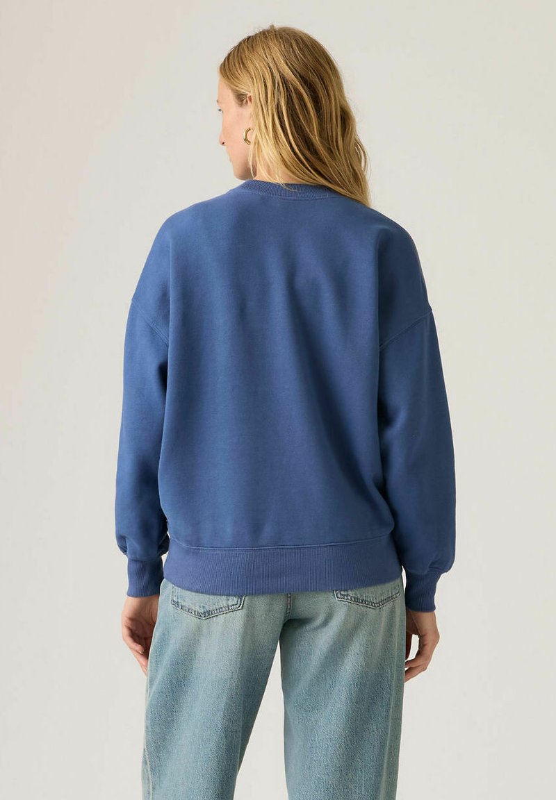 Femme aux longs cheveux blonds portant un sweat-shirt bleu et un jean bleu clair, vue de dos sur un fond neutre uni.