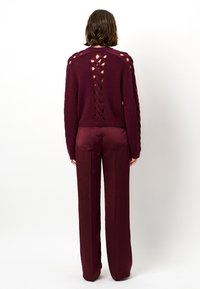 Bordeaux trui met uitsnijdingen aan de achterkant en mouwen, gecombineerd met bijpassende brede broek in een gladde, glanzende stof.
