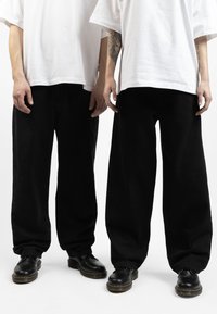 Pantaloni neri larghi con polsini elasticizzati, abbinati a t-shirt bianche oversize e scarpe nere con lacci e suola gialla.