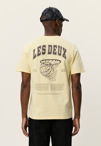 Κίτρινη κοντομάνικη μπλούζα με γραφικό ενός μπασκετικού διχτυού και κείμενο στην πλάτη. Κατασκευασμένη από βαμβάκι με άνετη εφαρμογή.