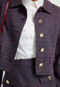 Giacca in tweed di un profondo blu e rosso con colletto, bottoni dorati e due tasche. Indossata sopra una blusa bianca con bottoni e dettagli in perle.