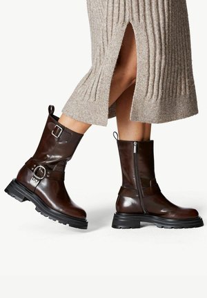 Bottes mi-mollet en cuir brun foncé avec boucles et fermetures éclair latérales portées avec une robe en maille côtelée beige avec une fente frontale.