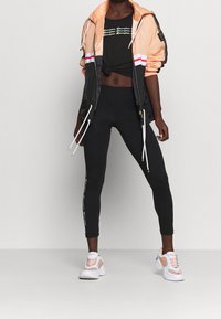 Mujer vistiendo leggings negros con logo lateral, camiseta corta negra de Champion, chaqueta de cremallera melocotón y negra, y zapatillas blancas con detalles melocotón.