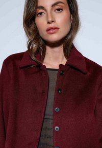 Bordeauxrode wollen mantel met een omgeslagen kraag, voorzien van zes zwarte knopen en een gladde textuur. Gedragen over een donkere top.