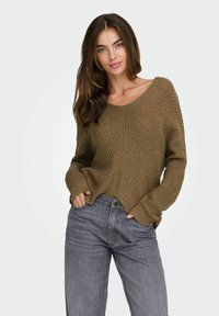 Maglione lavorato a maglia marrone con scollo a V e maniche lunghe, abbinato a jeans grigi. La texture del maglione appare morbida e a maglia allentata.