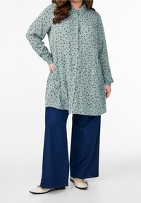 Camicia verde chiaro con bottoni e motivo floreale, abbinata a jeans a gamba larga blu e scarpe bianche con decorazioni.