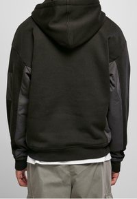 Persona con sudadera con capucha negra y paneles acolchados en las mangas, pantalones grises claros y una camiseta blanca visible debajo.