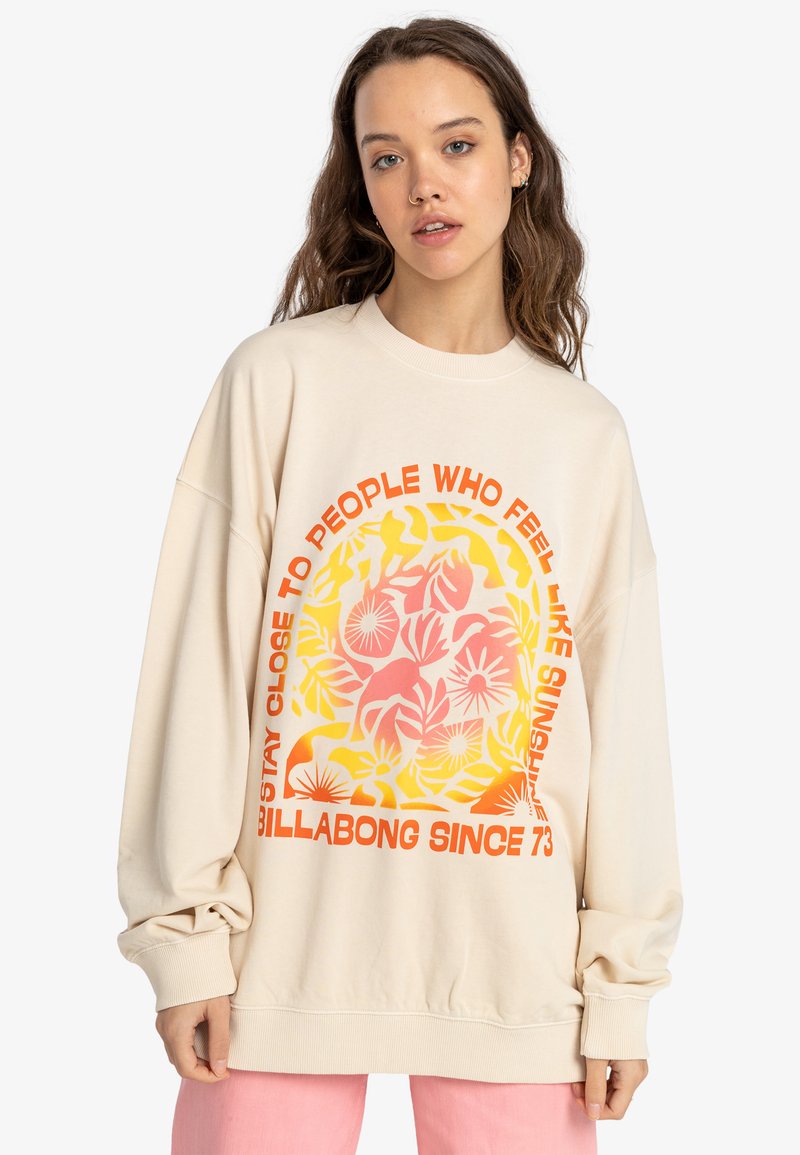 Oversized beige sweatshirt with colorful floral graphic and orange text. Features ribbed cuffs and hem, soft texture, and relaxed fit.