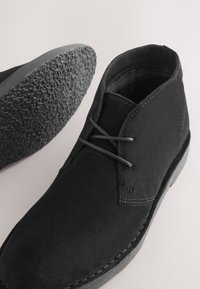 Bottines en daim noir avec semelles en caoutchouc texturées et lacets noirs, vues de dessus et de côté sur un fond clair.