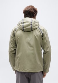 Άν δρας που φοράει αδιάβροχο μπουφάν ανοιχτού ελιάς με κουκούλα και σκούρο γκρι παντελόνι, απεικονισμένος από πίσω σε απλό φόντο.