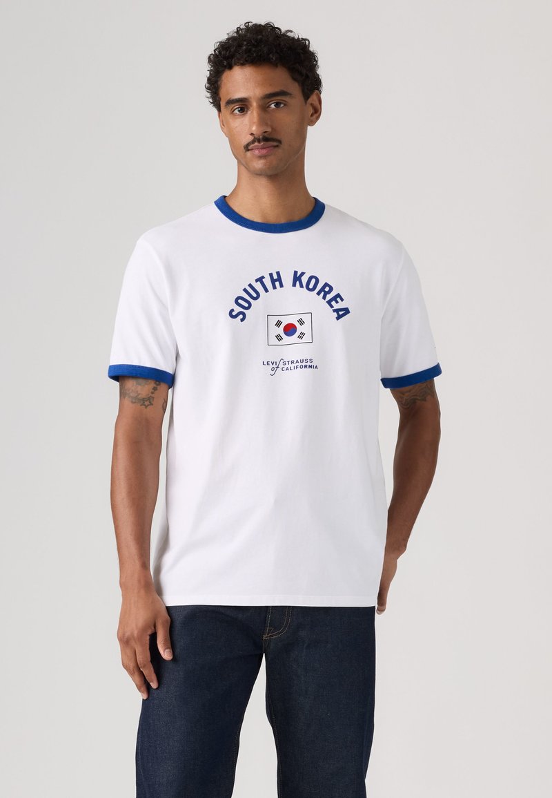 Homme portant un t-shirt blanc avec un liseré bleu, le drapeau de la Corée du Sud et un texte, associé à un jean en denim foncé, debout devant un fond uni.