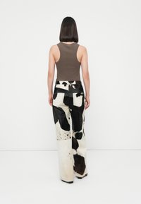 Jeans en fausse fourrure au motif vache, avec un design à jambe large, affichant un motif noir et blanc. Le mannequin porte un débardeur moulant marron.