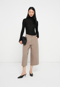 Czarny długi rękaw turtleneck, beżowe szerokie spodnie typu cropped, oraz czarne czółenka w szpic. Trzyma małą czarną torebkę.