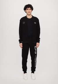 Black zip-up jacket with EA7 logo, paired with black sweatpants featuring white branding on the side, worn with white sneakers.