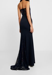 Granatowa suknia wieczorowa z elastycznego materiału, z koronkowym tyłem i trenem, charakteryzująca się dopasowanym krojem i bezramiennym fasonem.