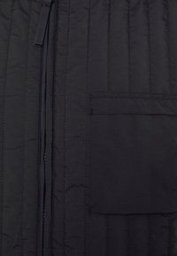 Svart quiltad puffjacka med vertikal sömnad, framdragkedja och en patchficka på vänster sida. Slät, matt textur.