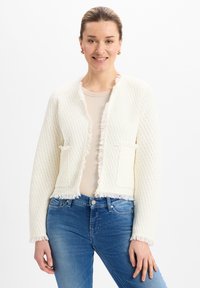 Cardigan bianco testurizzato con rifiniture in frange, apertura frontale e due tasche anteriori. Indossato sopra un top beige e abbinato a jeans blu.