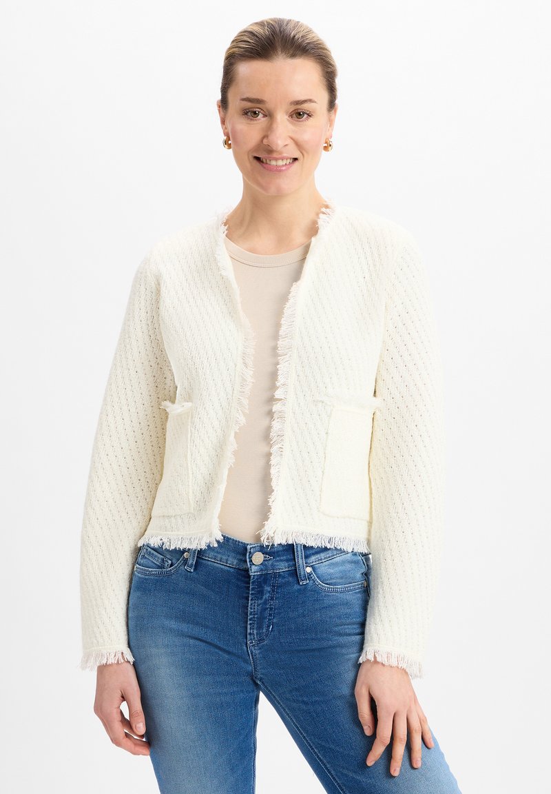 Cardigan bianco testurizzato con rifiniture in frange, apertura frontale e due tasche anteriori. Indossato sopra un top beige e abbinato a jeans blu.