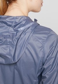Veste légère à capuche de couleur bleue douce, avec une texture lisse et brillante, et un cordon ajustable à la capuche pour un ajustement optimal.