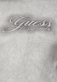 Pelēka ribota auduma virsma ar baltu "Guess Originals" logotipu kursīvā fontā. Gluda tekstūra ar vieglu piesātinājumu ap logotipu.